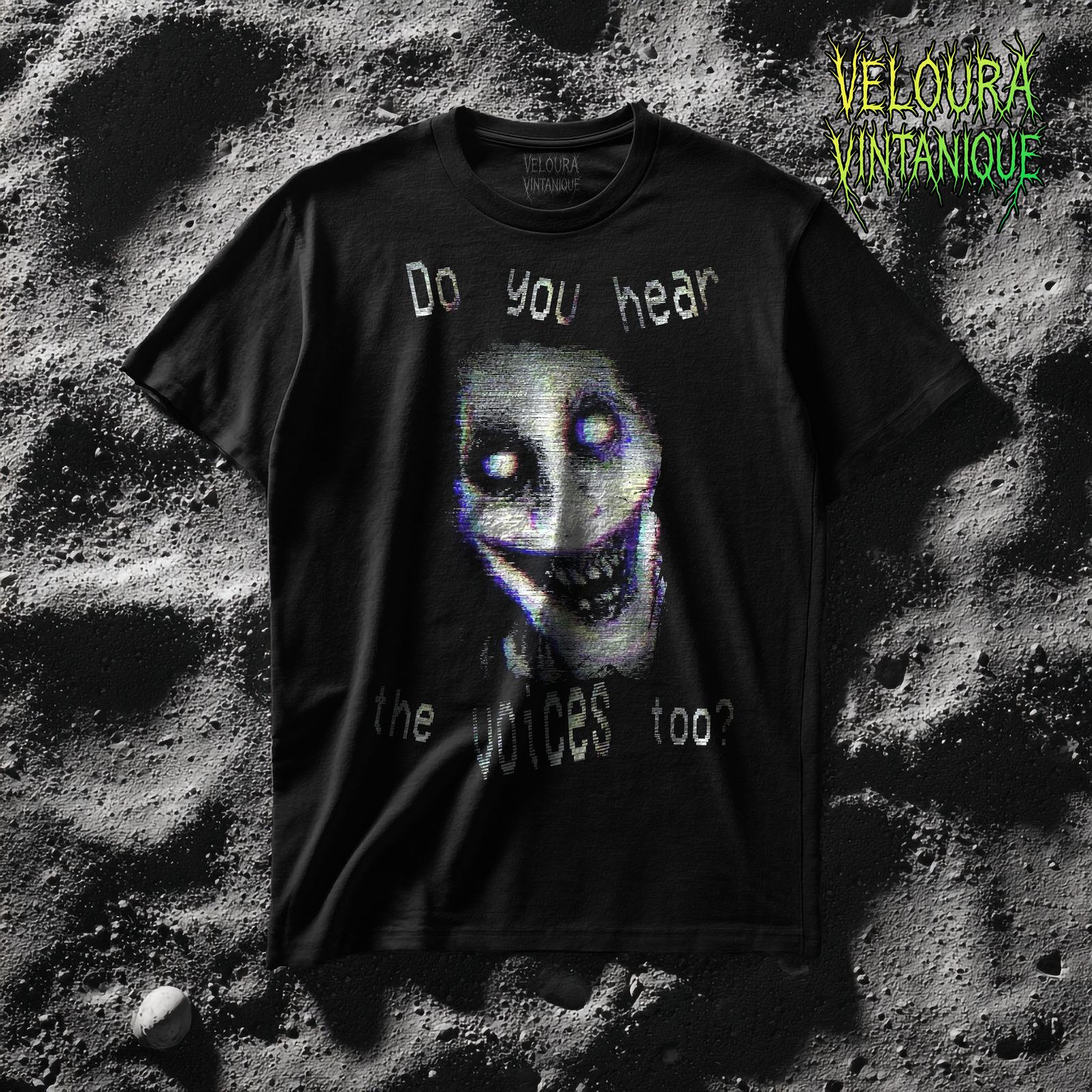 This t-shirt features a disturbing analog horror-inspired design. At the center, a twisted, grinning face with glowing white eyes and sharp, uneven teeth is rendered with a glitchy VHS static effect in eerie tones of white, purple, and blue. Surrounding the face, distorted text in a warped, pixelated font reads: “Do you hear the voices too?” giving the design a chilling psychological horror aesthetic.