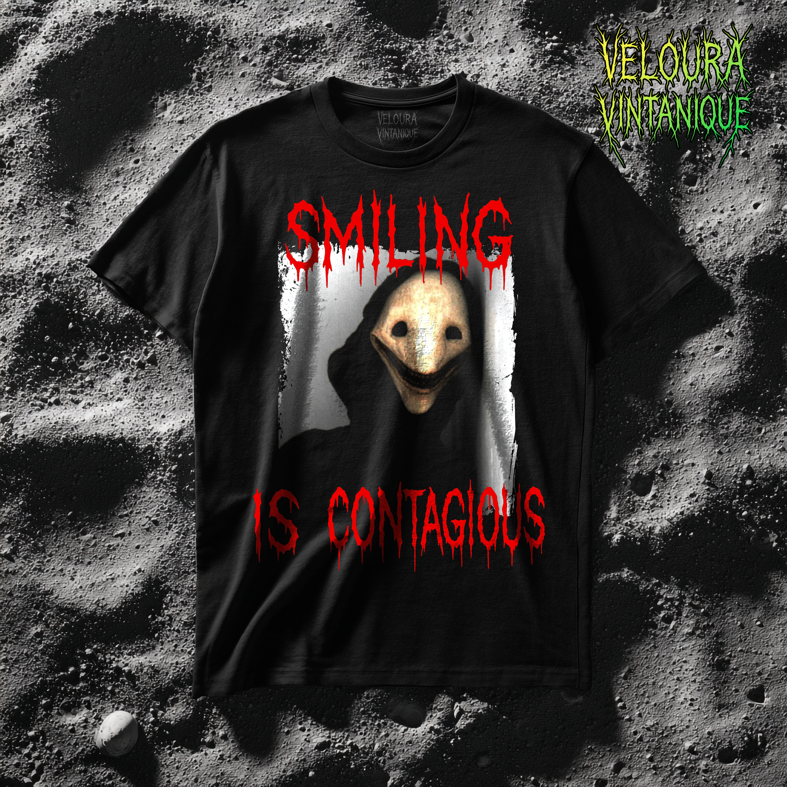 This horror t-shirt features a hooded figure with a pale mask-like face, hollow black eyes, and a wide unsettling grin, framed by the chilling phrase “SMILING IS CONTAGIOUS” in dripping blood-red letters above and below, creating a disturbing analog horror design with a creepy, nightmarish atmosphere.