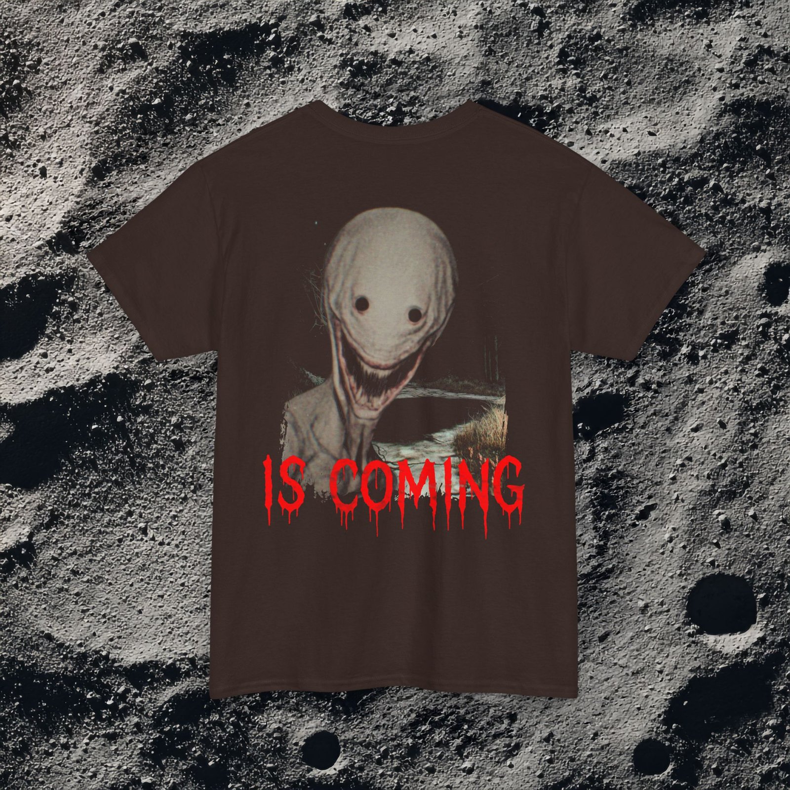 The shirt features a dark, desaturated forest path surrounded by dead trees and heavy shadows, creating a cold, abandoned atmosphere. Above the scene, dripping red text reads “THE FOG,” while on the opposite side a distorted, pale humanoid creature with hollow eyes and a wide, unnatural smile emerges from the misty background. Beneath it, the matching red text “IS COMING” completes the ominous analog-horror design.