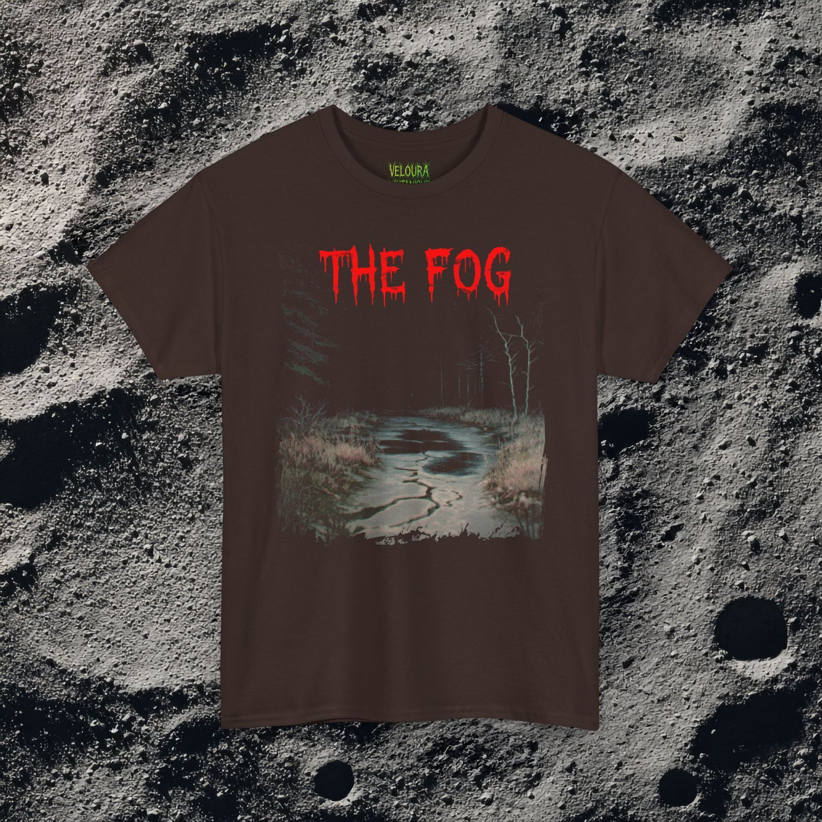 The shirt features a dark, desaturated forest path surrounded by dead trees and heavy shadows, creating a cold, abandoned atmosphere. Above the scene, dripping red text reads “THE FOG,” while on the opposite side a distorted, pale humanoid creature with hollow eyes and a wide, unnatural smile emerges from the misty background. Beneath it, the matching red text “IS COMING” completes the ominous analog-horror design.