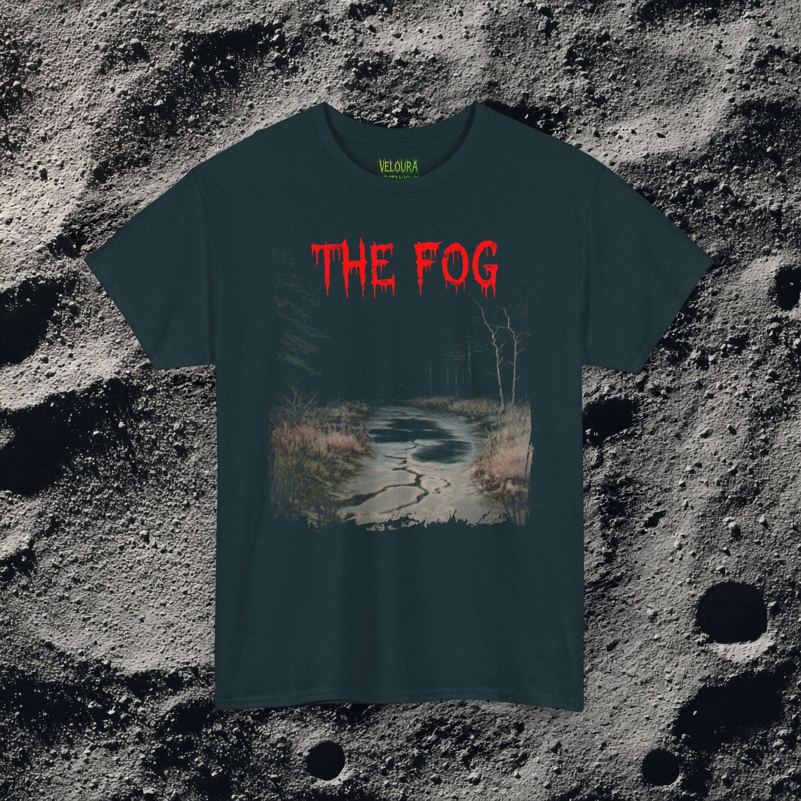 The shirt features a dark, desaturated forest path surrounded by dead trees and heavy shadows, creating a cold, abandoned atmosphere. Above the scene, dripping red text reads “THE FOG,” while on the opposite side a distorted, pale humanoid creature with hollow eyes and a wide, unnatural smile emerges from the misty background. Beneath it, the matching red text “IS COMING” completes the ominous analog-horror design.