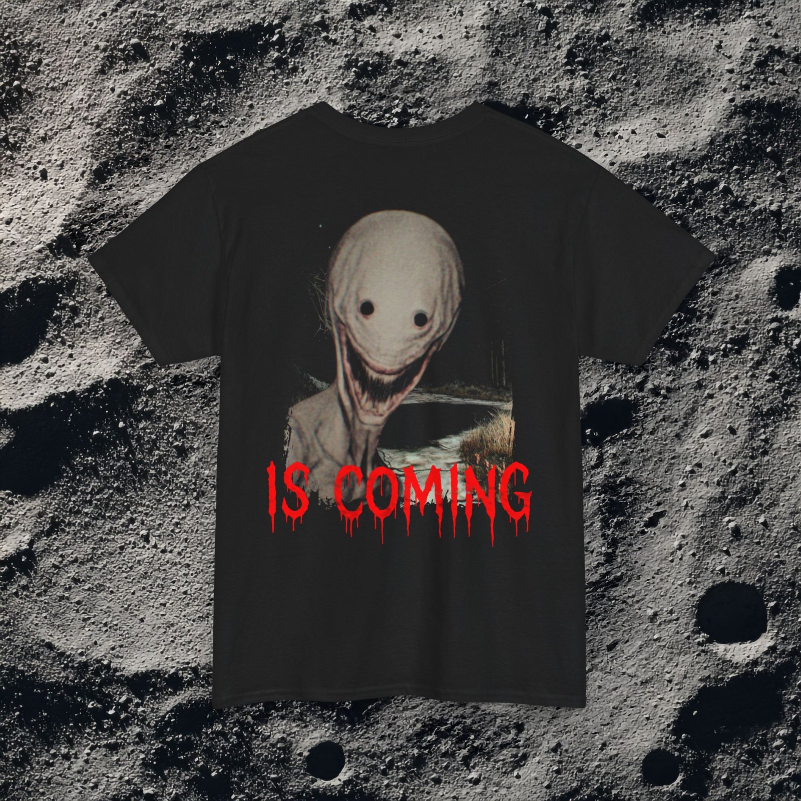 The shirt features a dark, desaturated forest path surrounded by dead trees and heavy shadows, creating a cold, abandoned atmosphere. Above the scene, dripping red text reads “THE FOG,” while on the opposite side a distorted, pale humanoid creature with hollow eyes and a wide, unnatural smile emerges from the misty background. Beneath it, the matching red text “IS COMING” completes the ominous analog-horror design.