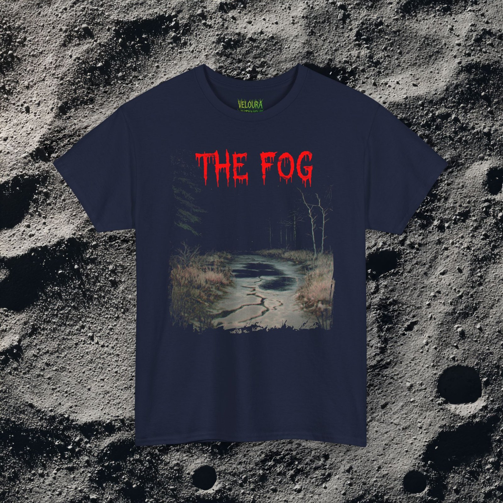 The shirt features a dark, desaturated forest path surrounded by dead trees and heavy shadows, creating a cold, abandoned atmosphere. Above the scene, dripping red text reads “THE FOG,” while on the opposite side a distorted, pale humanoid creature with hollow eyes and a wide, unnatural smile emerges from the misty background. Beneath it, the matching red text “IS COMING” completes the ominous analog-horror design.