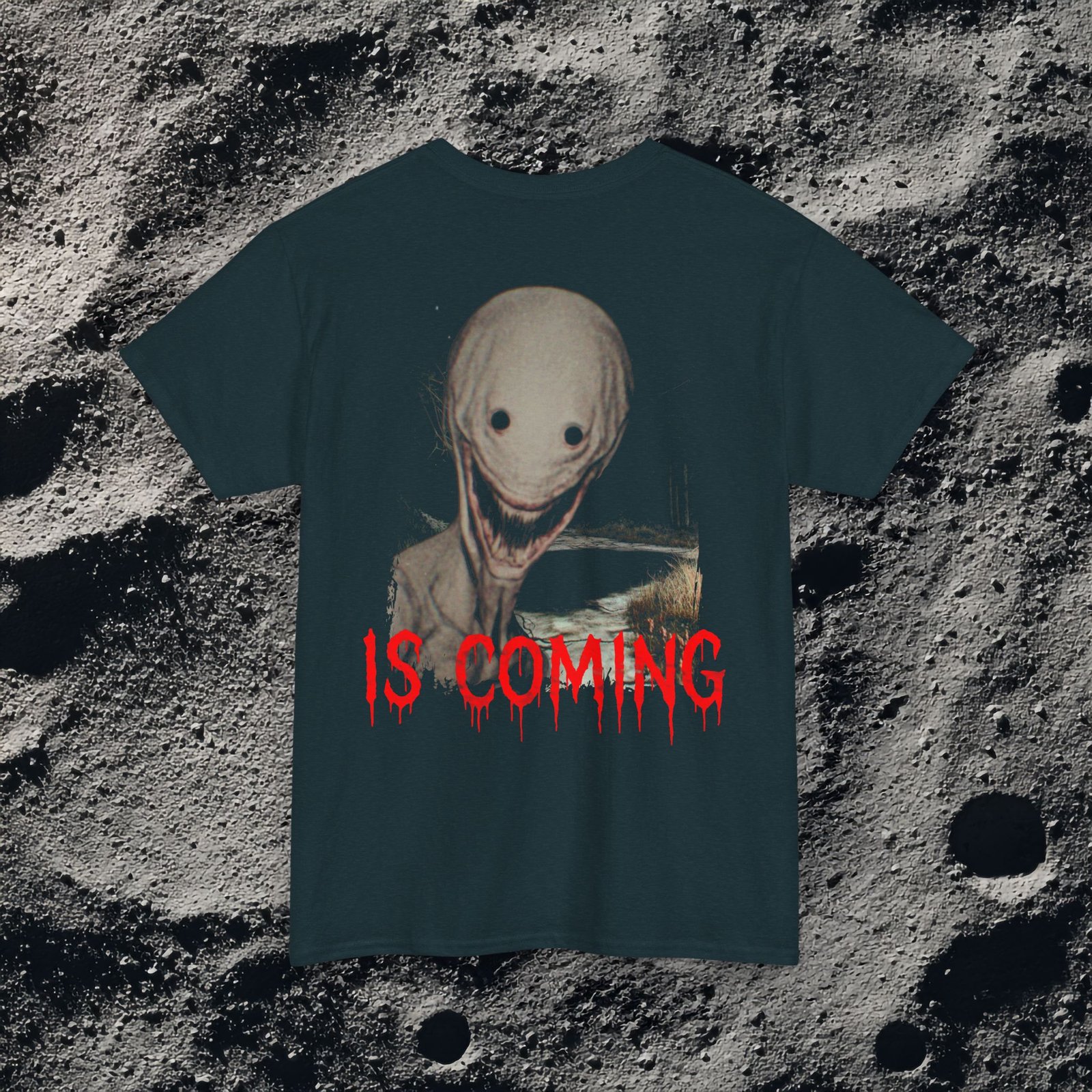 The shirt features a dark, desaturated forest path surrounded by dead trees and heavy shadows, creating a cold, abandoned atmosphere. Above the scene, dripping red text reads “THE FOG,” while on the opposite side a distorted, pale humanoid creature with hollow eyes and a wide, unnatural smile emerges from the misty background. Beneath it, the matching red text “IS COMING” completes the ominous analog-horror design.