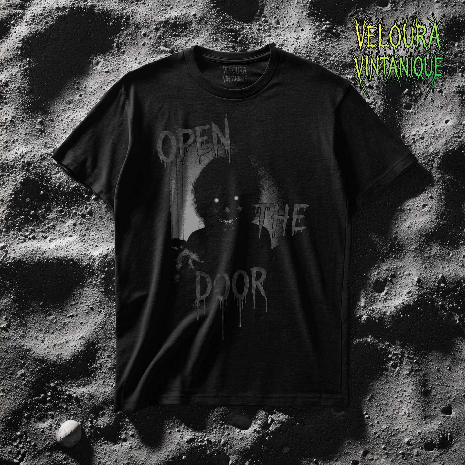 The shirt features a disturbing grayscale design of a shadowy humanoid creature emerging from a doorway, its glowing white eyes and jagged grin piercing through the darkness, with its faintly visible claw-like hands reaching forward, surrounded by heavy film grain and harsh lighting; above, beside, and below the creature are dripping horror-style letters forming the phrase “OPEN THE DOOR,” the text appearing smeared, melting, and integrated into the eerie atmosphere of the image.